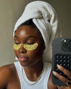 Mulher jovem de pele escura com toalha de banho na cabeça e máscara para os olhos.