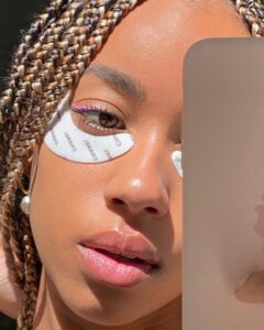 Mulher jovem de pele negra em foto no espelho com material de skin care no rosto.