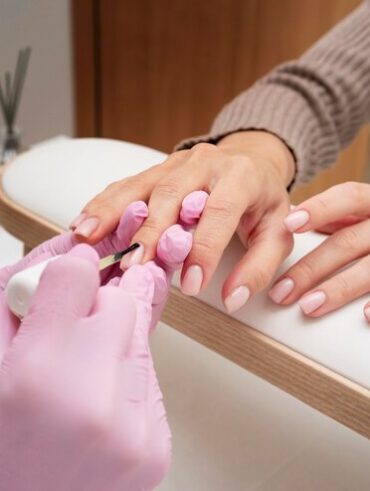 duas mãos com luvas rosas e acessórios de manicure e duas mãos da cliente que está fazendo a unha