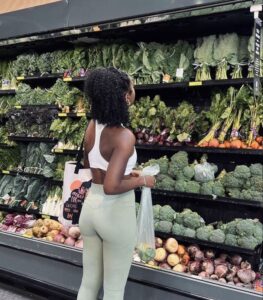 Mulher jovem e magra com roupa de academia na frente de uma geladeira de mercado com salada verde