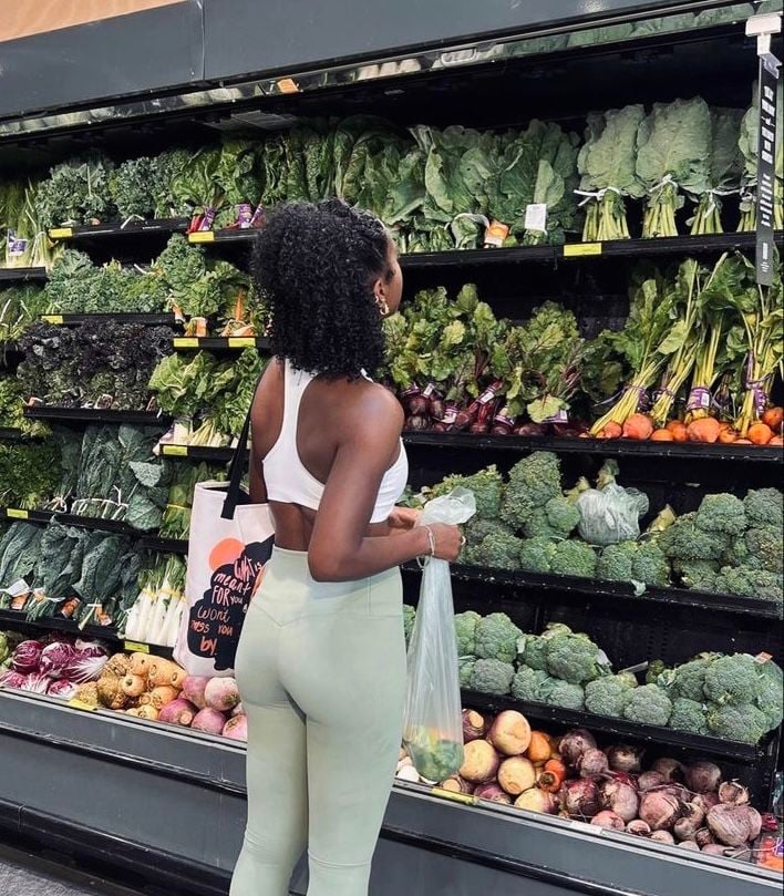 Mulher jovem e magra com roupa de academia na frente de uma geladeira de mercado com salada verde
