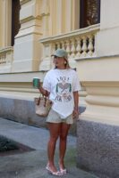 Mulher jovem, loira e magra. Ela veste camiseta estampada branca, bermuda e salto alto.