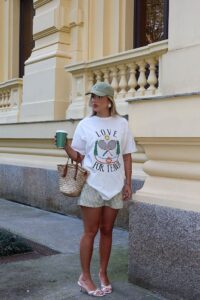 Mulher jovem, loira e magra. Ela veste camiseta estampada branca, bermuda e salto alto.