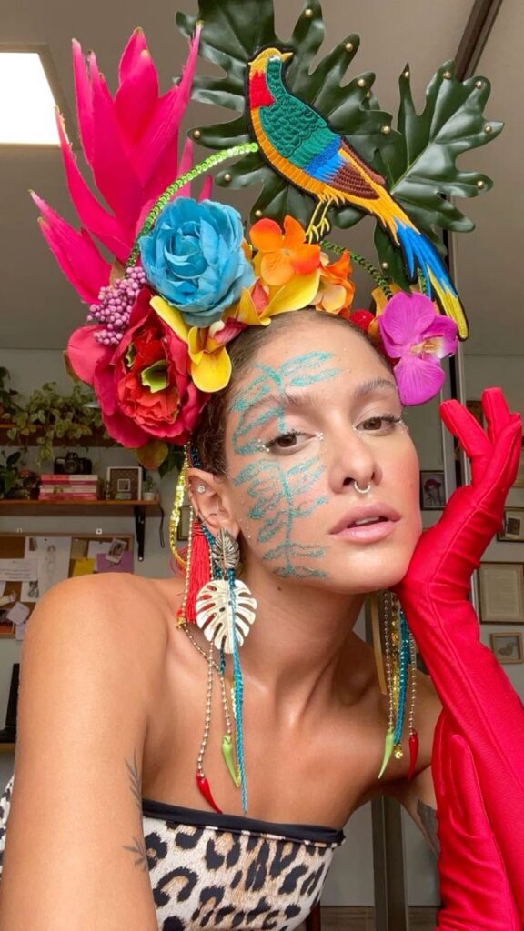 Mulher jovem de pele clara com look de carnaval. Ela usa tiara com flores, folhas e um pavão, brincos longos, luvas vermelhas e roupa com estampa de leopardo.
