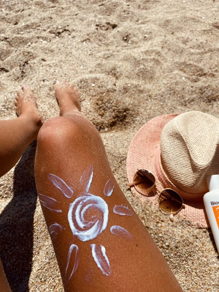 Perna de pele 'média, apoiada na areia, com chapéu e óculos no chão. Além disso, na perna tem um sol desenhado com protetor solar