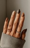 Mão de pele clara, com acessórios em prata e dourado e unha amendoada degradê em tons terrosos