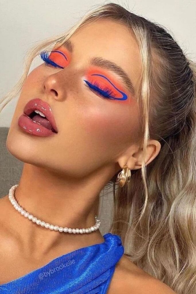 Brookelle McKenzie, com blusa azul, colar de pérola, cabelo loiro preso, e maquiagem com sombra laranja e delineado gráfico azul