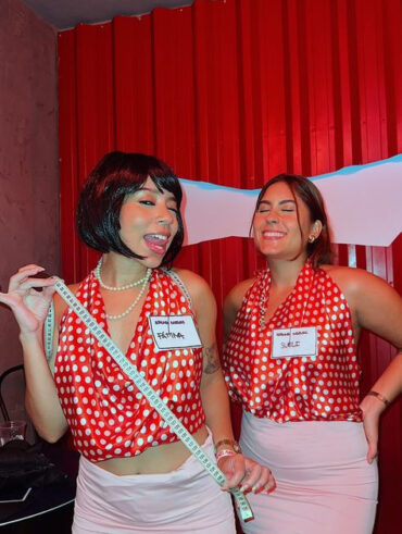 Duas mulheres de pele clara, com saia rosa-claro e blusa vermelha com bolinhas