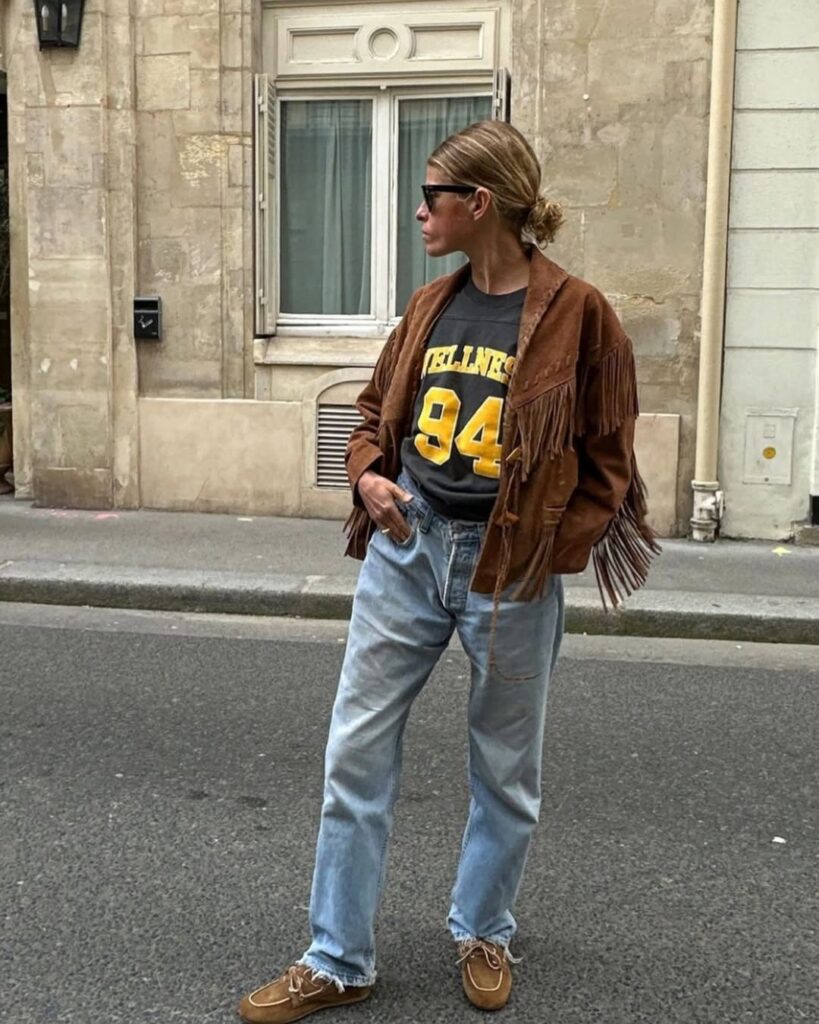 Mulher de pele clara, com calça jeans, camisa estampada, jaqueta de franja e calçado de camurça 