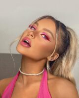 Brookelle, com cabelo loiro meio preso, top rosa, acessórios com pérolas e maquiagem com delineado rosa e vermelho