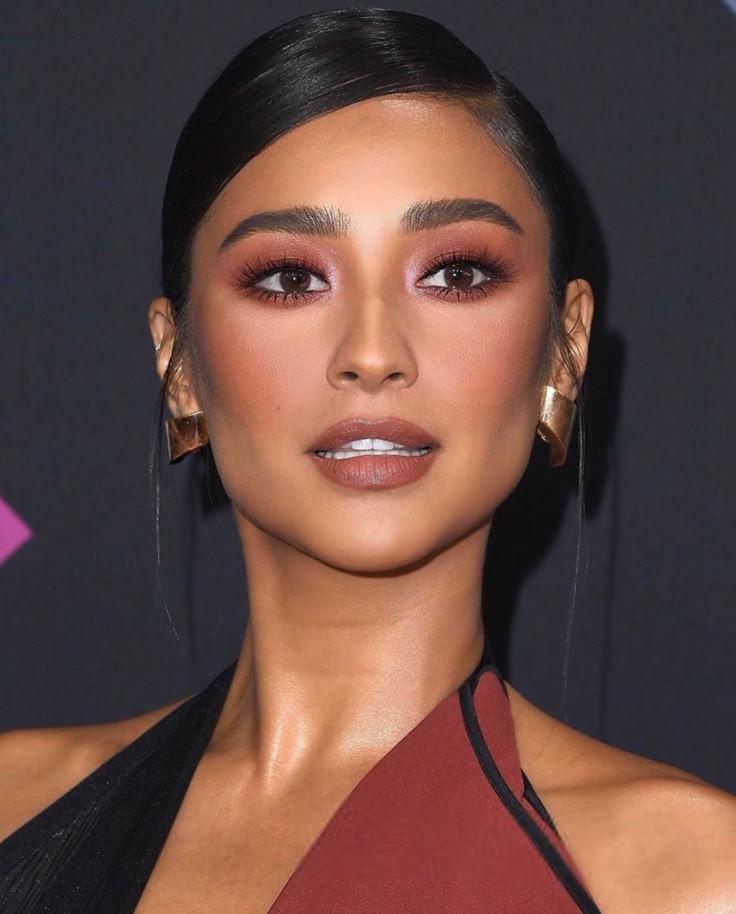 Shay Mitchell com vestido preto, cabelo escuro preso, acessórios em dourado e maquiagem em tons terrosos com batom marrom-claro 