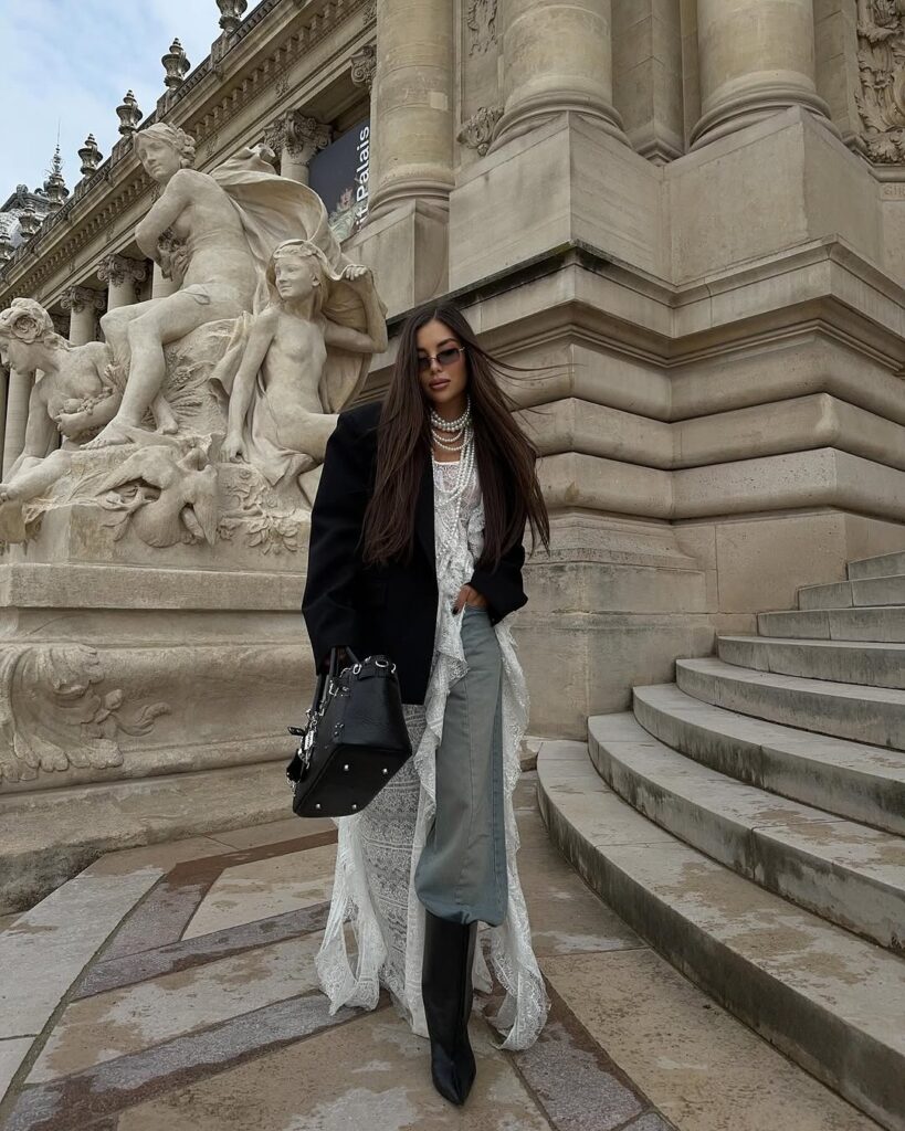 Karina Nigay, com blusa de renda, calça jeans, casaco, bota e bolsa preta e colar de pérolas