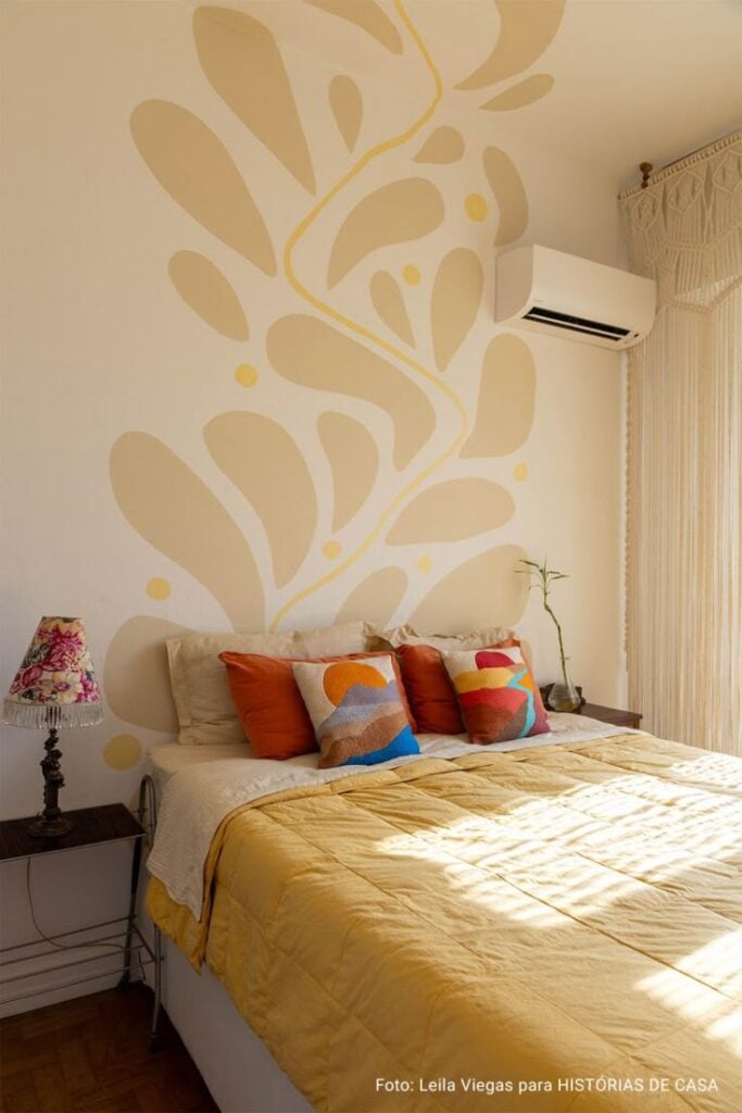 Quarto com parede bege e pintura orgânica em tom neutro, edredom amarelo e almofadas coloridas