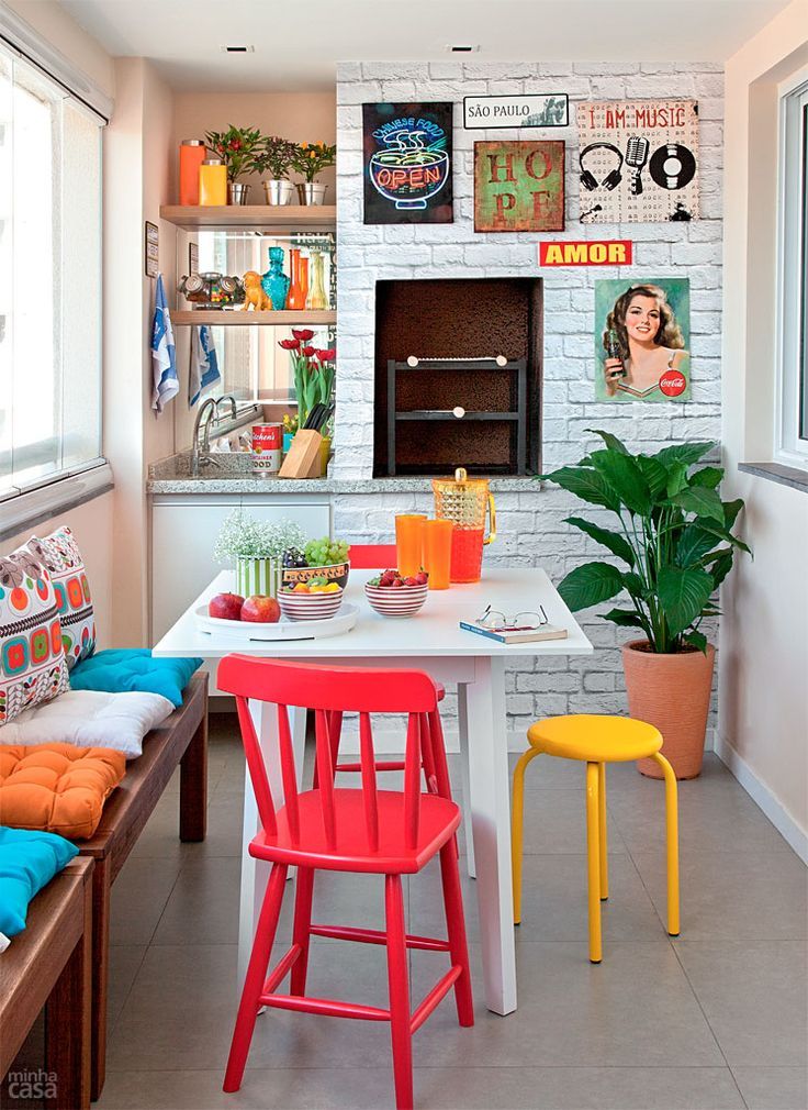 Cozinha com papel de parede de tijolinhos brancos com quadros, mesa branca e cadeiras coloridas