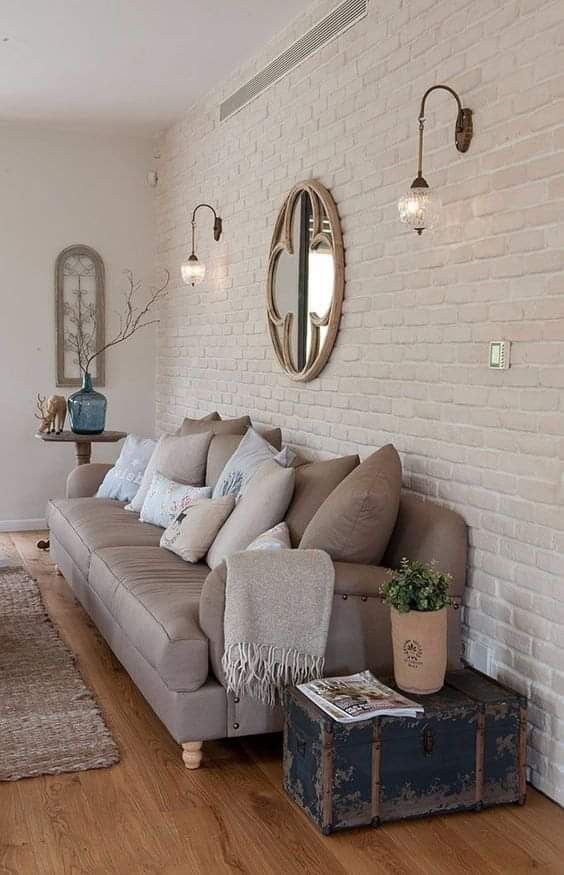 Sala com tijolinhos brancos, sofá marrom, espelho redondo e arandelas
