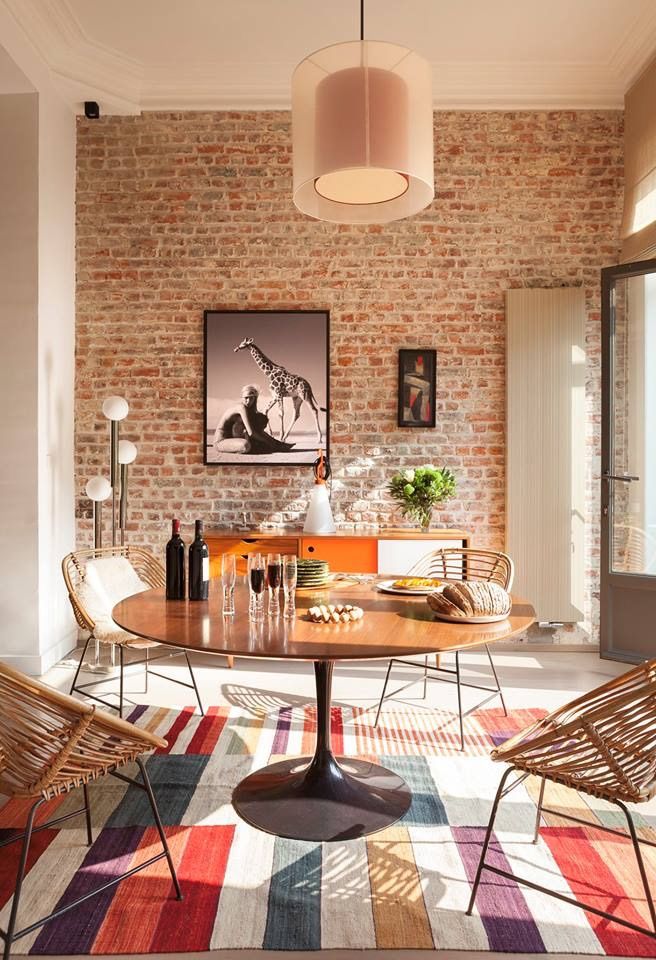 Sala de jantar com mesa redonda, tapete com listras coloridas, parede de tijolinhos e quadros