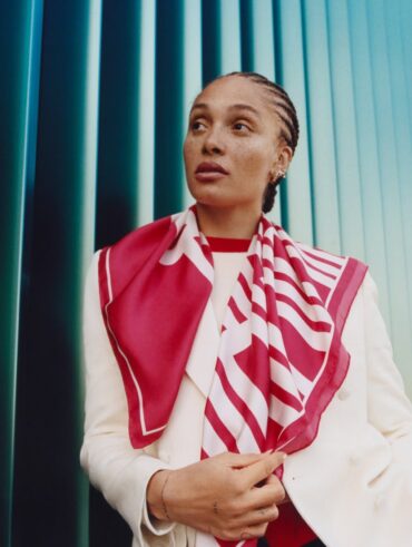 Mulher jovem e magra de pele escura. Ela usa blazer branco e lenço branco com vermelho.