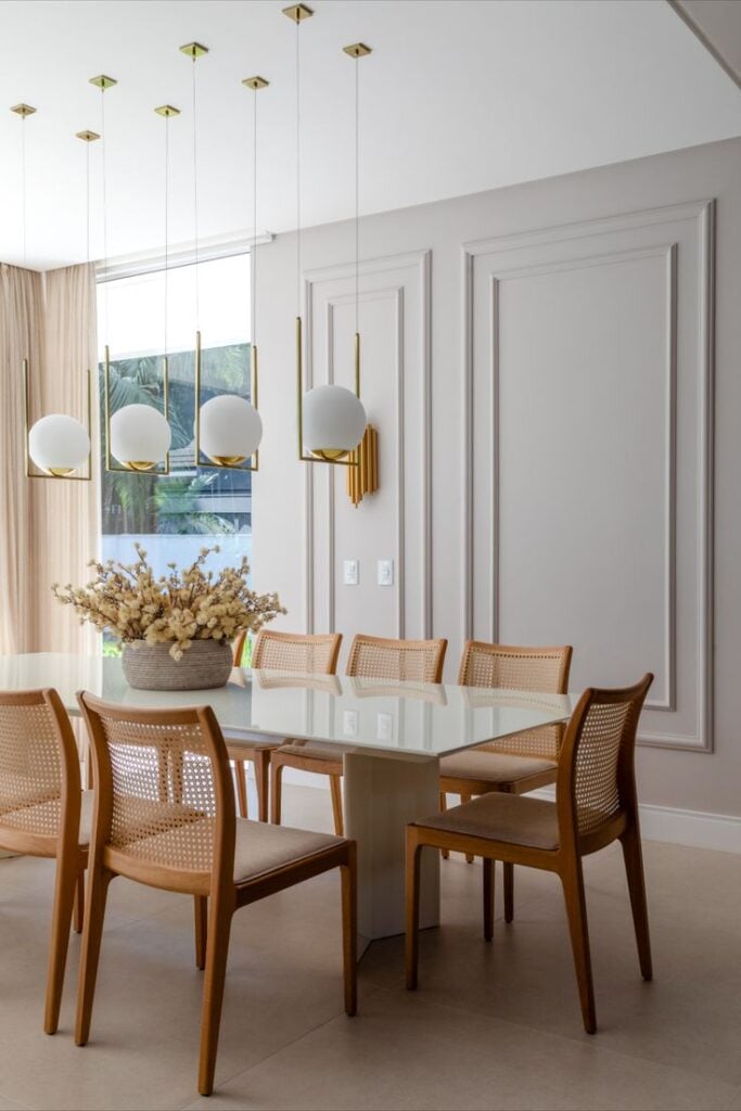 Sala de jantar neutra, com cadeiras em madeira com encosto de palhinha, parede com boiseries e pendentes