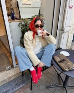 Mulher jovem sentada em uma cadeira na frente de um café. Ela usa casaco, blusa e calça jeans. Além disso, ela está usando o tenis da Puma speedcat em modelo vermelho, e um lenço vermelho na cabeça.