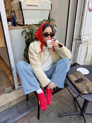 Mulher jovem sentada em uma cadeira na frente de um café. Ela usa casaco, blusa e calça jeans. Além disso, ela está usando o tenis da Puma speedcat em modelo vermelho, e um lenço vermelho na cabeça.