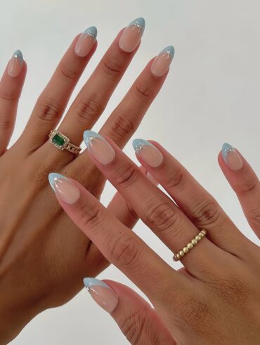 Mãos de pele clara, com anéis em dourado e unha amendoada nude com francesinha azul Tiffany com laço