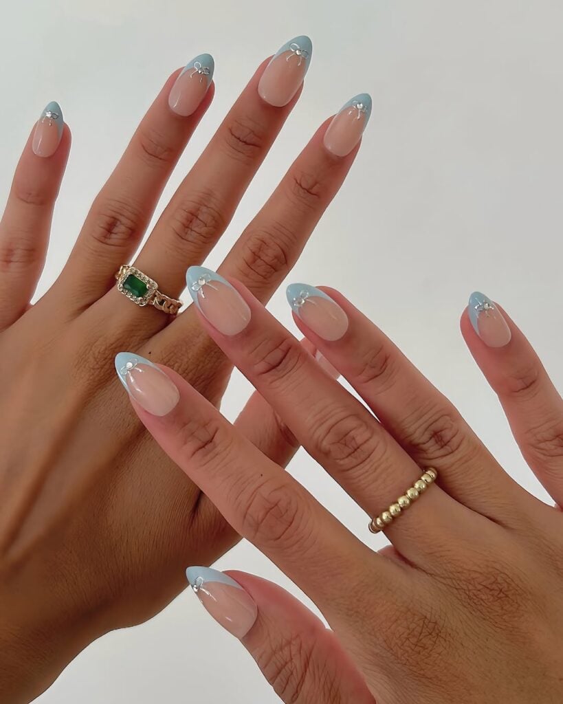 Mãos de pele clara, com anéis em dourado e unha amendoada nude com francesinha azul Tiffany com laço 