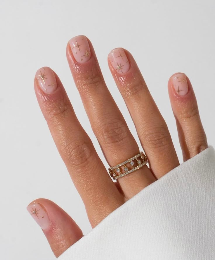 Mão de pele clara, com anel de brilhante dourado e unha curta nude e decorações com estrelas em dourado