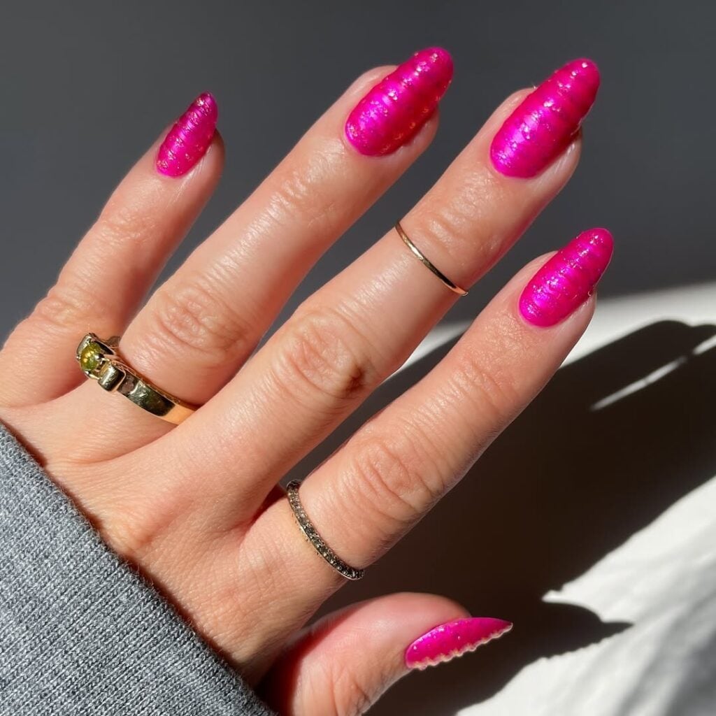 Mão de pele clara, com anéis em dourado e unha amendoada hot pink com decorações com glitter