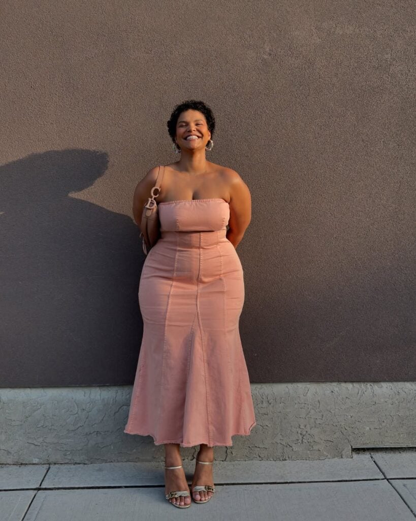 Mulher de pele preta e midsize, com vestido rosa-antigo sem alça 