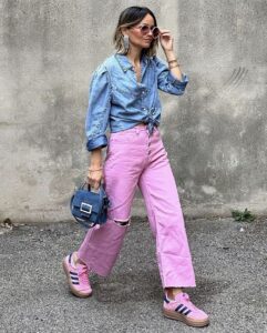 Mulher jovem, magra e de pele clara. Ela usa camisa jeans, calça jeans e tênis Adidas