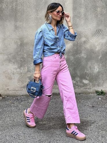 Mulher jovem, magra e de pele clara. Ela usa camisa jeans, calça jeans e tênis Adidas