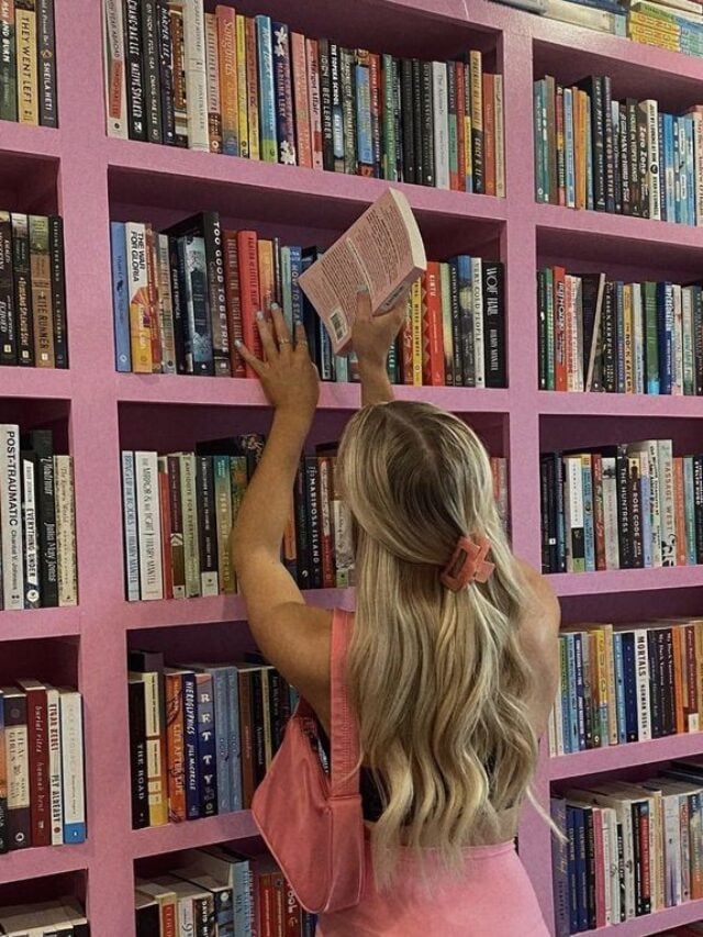 Mulher loira e de pele clara, com look rosa em uma livraria