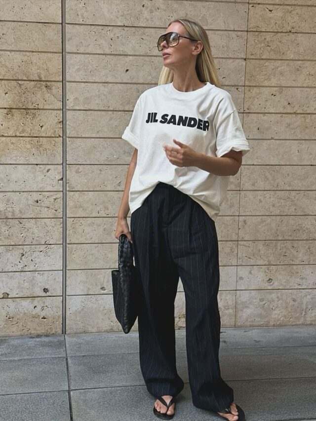 Mulher magra e de pele clara, com calça de alfaiataria, blusa oversized, bolsa e chinelo preto
