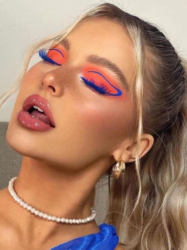 Brookelle McKenzie, com blusa azul, colar de pérola, cabelo loiro preso, e maquiagem com sombra laranja e delineado gráfico azul
