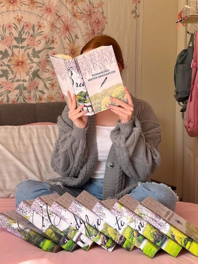 Mulher de ele clara, com cabelo ruivo preso, calça jeans, blusa branca e casaquinho cinza, lendo um livro de Bridgerton