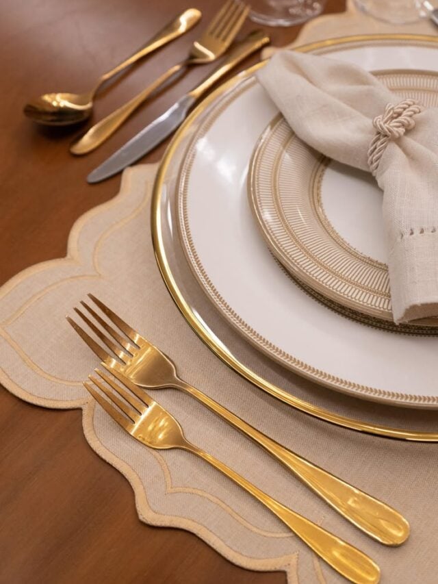 Mesa com jogo americano bege, prato com borda dourada e talheres dourados