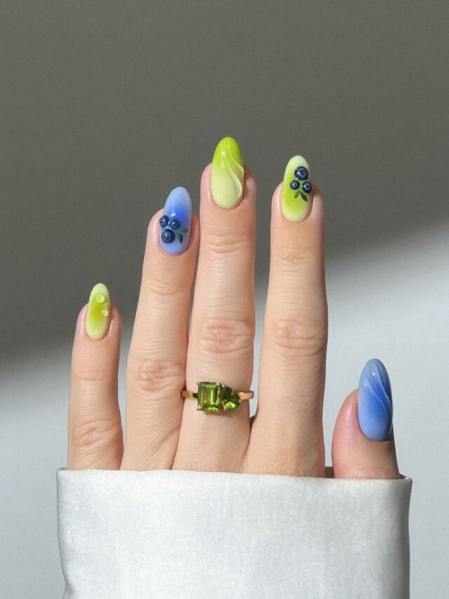 Mão de pele clara, com anel verde e unha amendoada verde e azul com decoração em gel e de fruta
