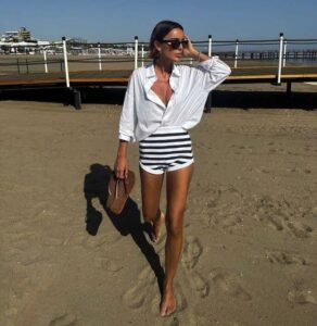 mulher jovem com look estilo fisherman core. Ela usa camisa branca, hot pants branco com listras azuis e óculos de sol