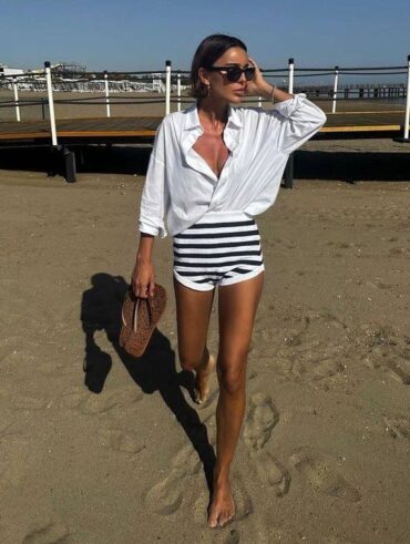 mulher jovem com look estilo fisherman core. Ela usa camisa branca, hot pants branco com listras azuis e óculos de sol