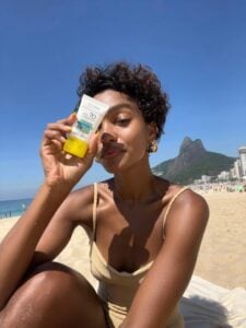 Mulher jovem de pele negra e cabelos curtos e escuros. Ela está na beira da praia e veste maiô e segura um protetor solar na frente do rosto.