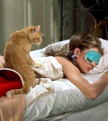 Mulher jovem deitada em uma cama com máscara de descanso para os olhos. Em suas costas, está um gatinho laranja.
