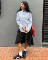Mulher jovem de pele escura e cabelos escuros. Ela usa camisa de manga longa e saia, junto de um tenis speedcat preto da Puma.