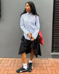 Mulher jovem de pele escura e cabelos escuros. Ela usa camisa de manga longa e saia, junto de um tenis speedcat preto da Puma.