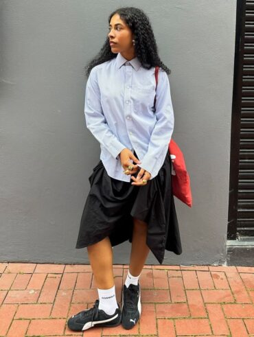 Mulher jovem de pele escura e cabelos escuros. Ela usa camisa de manga longa e saia, junto de um tenis speedcat preto da Puma.