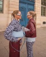 Duas mulheres de pele clara. A mais próxima da câmera está usando camisa de zebra, calça bordô e acessórios em azul claro. A outra, está vestindo o contrário