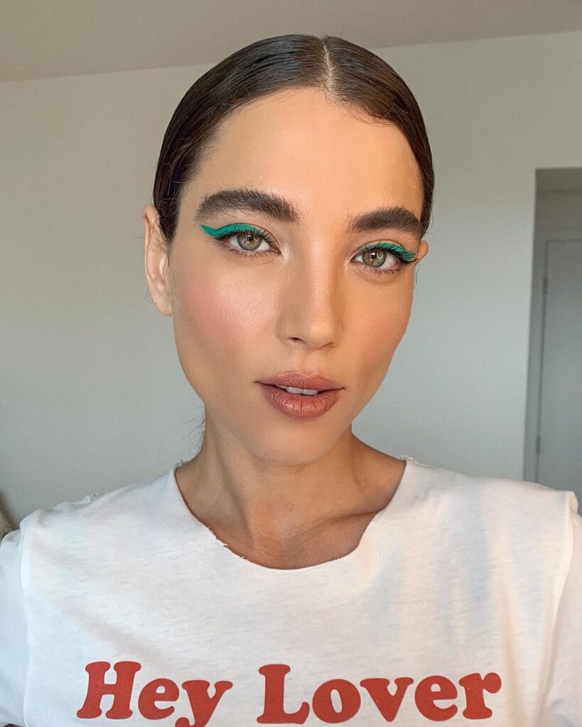 Mulher de pele clara, com blusa branca, cabelo escuro preto e maquiagem com delineado azul-esverdeado 