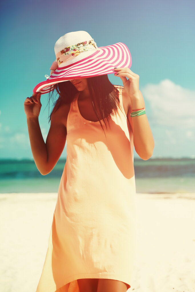 Mulher na praia com vestido amarelo e chapéu com aba listrada em vermelho e branco