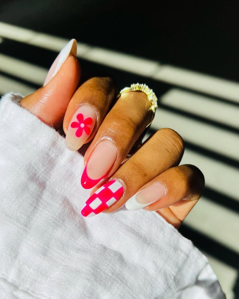 Mão de pele preta, com anel em dourado e unha amendoada com francesinha em branco e rosa. Além disso, também há decoração com flores e quadriculado 