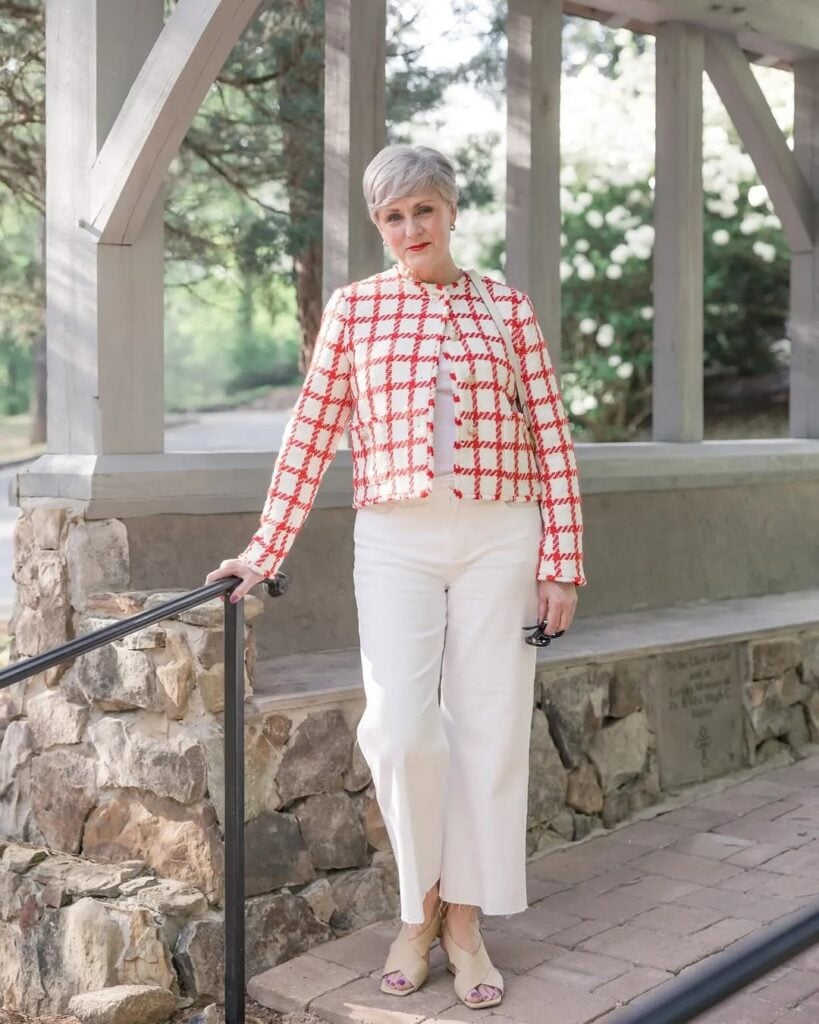 Mulher com mais de cinquenta anos, pele clara, cabelos curtos e grisalhos. Ela usa casaco listrado branco e vermelho, calça jeans branca e tamanco bege.