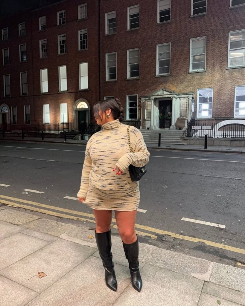 Mulher jovem com cabelo escuro. Ela usa vestido de manga longa curto e justo e botas pretas de cano longo